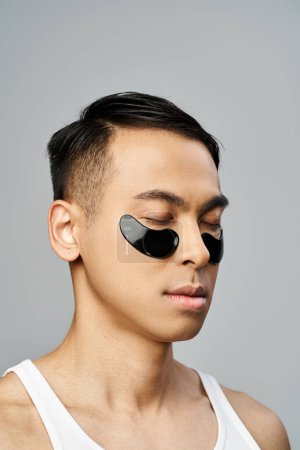 Hombre asiático guapo con un parche de ojo negro durante la rutina de belleza en el estudio gris.