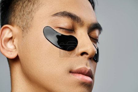 Handsome Asian man in a beauty routine, wearing a black eye patch in a grey studio setting.