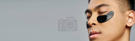 Foto de Hombre asiático guapo luciendo un parche en un estudio gris durante una rutina de belleza y cuidado de la piel. - Imagen libre de derechos