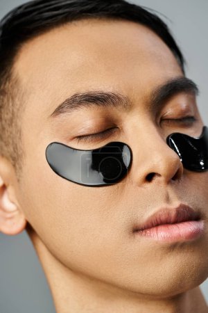 Bel homme asiatique subissant une routine beauté et soins de la peau, portant des bandeaux oculaires noirs dans un cadre de studio gris.