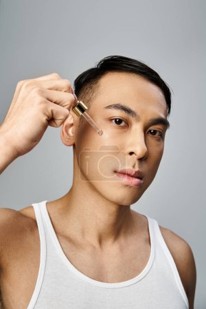 A handsome Asian man using serum during a beauty routine in a grey studio setting.