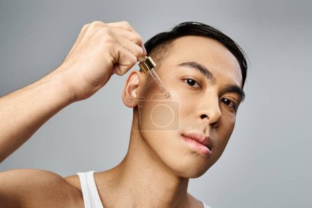 Un hombre asiático guapo sosteniendo pipeta con suero cerca de la mejilla durante una rutina de belleza y cuidado de la piel en un estudio gris.
