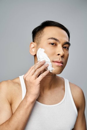 Schöner asiatischer Mann rasiert sich sanft sein Gesicht, die Augen fokussiert in einem grauen Studio-Setting.