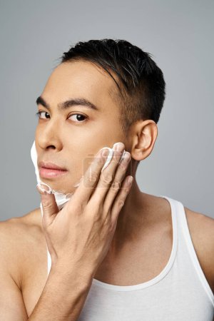 A handsome Asian man with shaving foam on his face in a grey studio.
