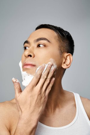 Guapo, hombre asiático con espuma de afeitar en la cara, en un entorno de estudio gris.