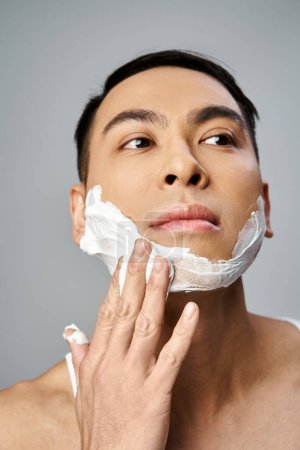 Hombre asiático guapo con espuma de afeitar en la cara en un estudio gris.