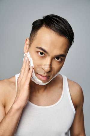 Hombre asiático guapo con espuma de afeitar en la cara, cortésmente afeitado en un estudio gris.