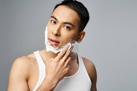 Hombre asiático guapo con espuma de afeitar en la cara, afeitándose meticulosamente con una navaja en un estudio gris.