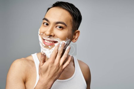 A handsome Asian man with shaving foam on face, meticulously shaving with a razor in a grey studio.
