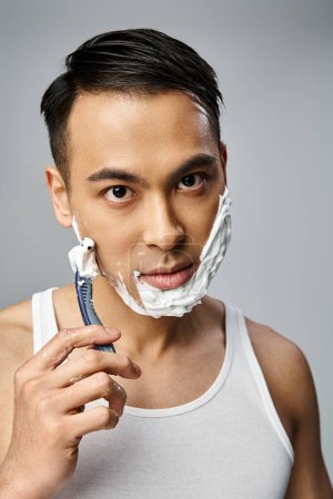 An Asian man with shaving foam on his face attentively shaves with a razor in a serene grey studio setting.