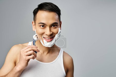Ein hübscher asiatischer Mann mit Rasierschaum im Gesicht rasiert sich in einem grauen Studio vorsichtig mit einem Rasiermesser.
