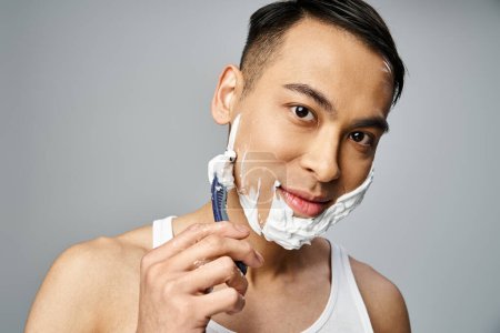 Ein hübscher asiatischer Mann mit Rasierschaum im Gesicht, der in einem grauen Studio ein Rasiermesser benutzt.