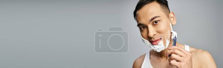 Téléchargez les photos : Un homme asiatique avec de la mousse à raser sur son visage se rasant soigneusement les cheveux du visage avec un rasoir dans un studio gris. - en image libre de droit