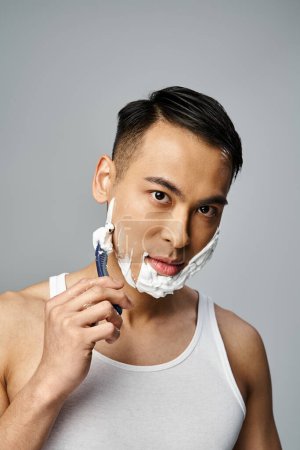 A stylish Asian man in black attire gracefully shaves his face with a razor in a vibrant red studio setting.