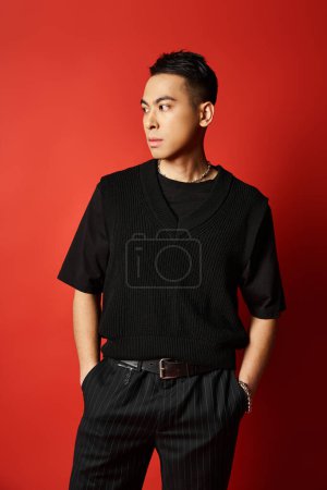 A stylish and handsome Asian man dressed in black stands confidently in front of a bold red wall in a studio setting.