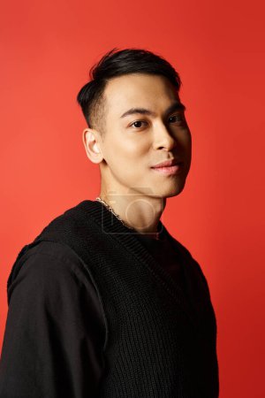 Handsome Asian man exudes charm in a black, standing confidently against a striking red backdrop in a studio setting.