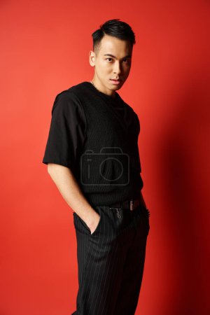 A stylish Asian man in black attire confidently stands in front of a vibrant red wall in a studio setting.