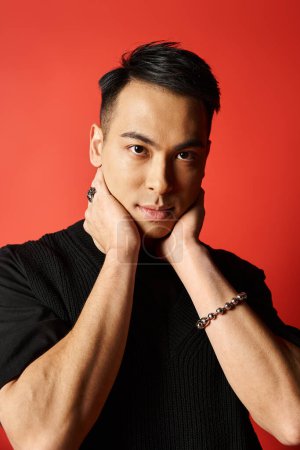 A stylish Asian man in black attire strikes a pose with his hand on his chin against a vibrant red background in a studio setting.
