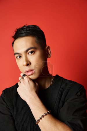 A stylish Asian man in black attire strikes a pose with hand on chin against a vibrant red backdrop in a studio setting.