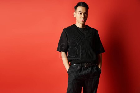 Téléchargez les photos : Un homme asiatique élégant et beau en tenue noire pose devant un mur rouge audacieux dans un décor de studio. - en image libre de droit