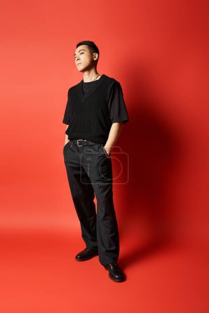 A fashionable Asian man in black attire stands confidently in front of a striking red wall in a studio setting.