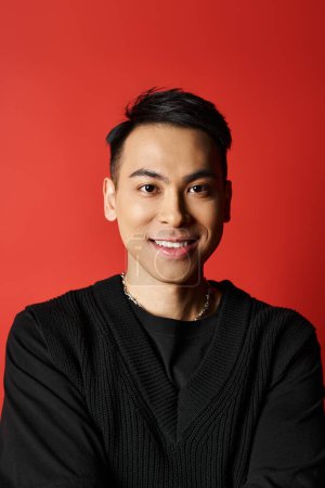 Ein gut aussehender asiatischer Mann lächelt freundlich in die Kamera, trägt einen schicken schwarzen Pullover vor leuchtend roter Studiokulisse.