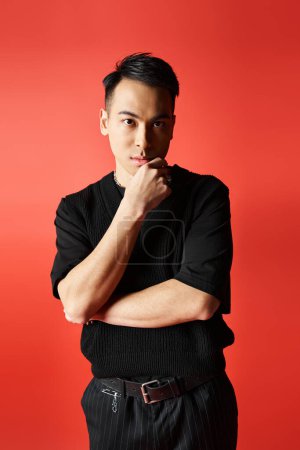 Foto de Elegante y guapo hombre asiático vestido de negro apoyando su barbilla en su mano, profundamente en pensamiento sobre un fondo rojo. - Imagen libre de derechos
