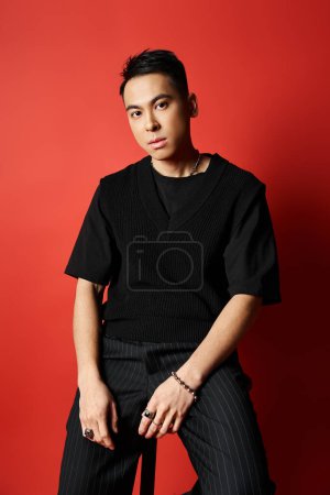 Téléchargez les photos : A stylish, handsome Asian man in a black shirt sits on a chair against a vibrant red background in a studio setting. - en image libre de droit