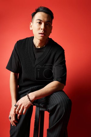 A stylish Asian man in black attire sits on a stool in front of a striking red wall in a studio setting.