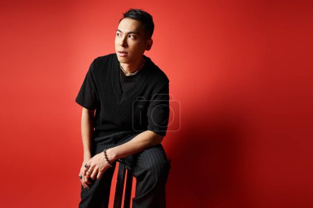 A stylish and handsome Asian man in black attire sits thoughtfully on a chair in front of a vibrant red wall in a studio setting.