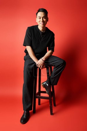 Téléchargez les photos : Un bel homme asiatique en tenue noire s'assoit élégamment sur un tabouret sur un fond rouge frappant. - en image libre de droit