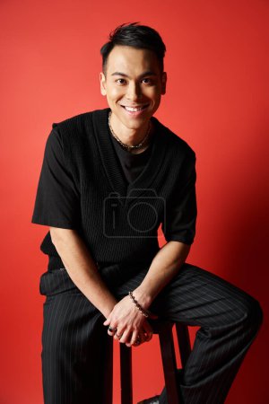 A stylish, handsome Asian man sits on a stool in front of a red wall, wearing black attire in a studio setting.