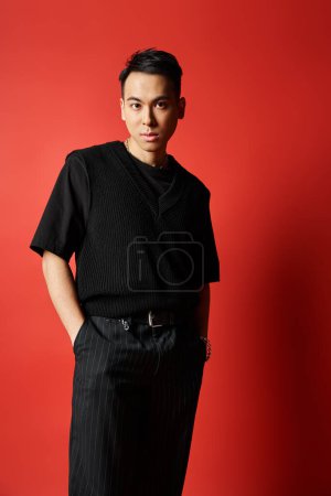 Ein stilvoller asiatischer Mann in schwarzer Kleidung steht selbstbewusst vor einer leuchtend roten Wand in einem Studio-Ambiente.
