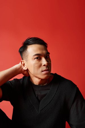 Photo for A stylish, handsome Asian man in a black sweater strikes a pose on a red background in a studio setting. - Royalty Free Image