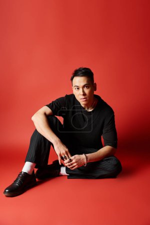 Stylish Asian man in black attire sitting on the ground with legs crossed in a zen pose on a vibrant red background.