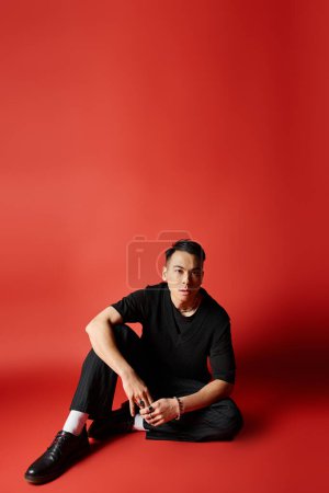 Foto de Un elegante hombre asiático vestido de negro se sienta con gracia en el suelo sobre un llamativo fondo rojo. - Imagen libre de derechos