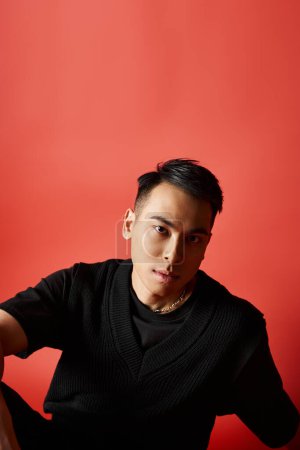 Ein stilvoller und gut aussehender asiatischer Mann in schwarzem Hemd steht selbstbewusst vor einer leuchtend roten Wand in einem Atelier.