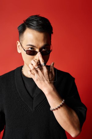 A stylish, handsome Asian man in a black attire holding a sunglasses against a bold red background in a studio setting.