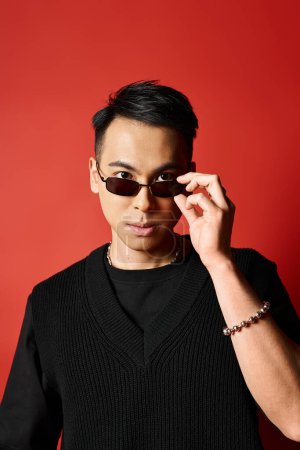 Stylish and handsome Asian man wearing a black sunglasses posing in a studio with a red background.