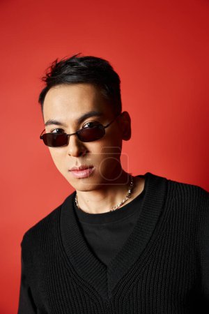 A handsome Asian man poses in a black outfit and sunglasses against a vibrant red backdrop.