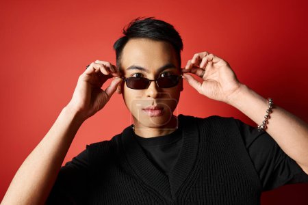 Téléchargez les photos : Un homme asiatique élégant en chemise noire et lunettes de soleil pose en toute confiance sur un fond rouge vif. - en image libre de droit