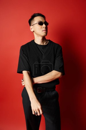 A stylish and handsome Asian man dressed in a black shirt and black pants poses against a bold red background in a studio.