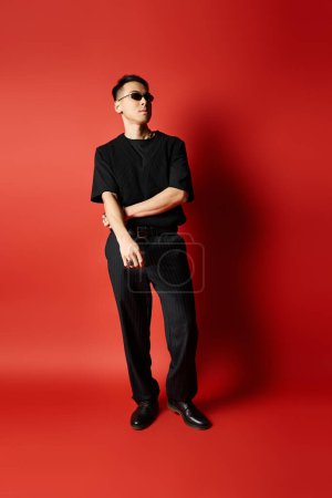 Ein stilvoller asiatischer Mann in schwarzer Kleidung steht selbstbewusst vor einem fetten roten Hintergrund in einem Studio-Setting.