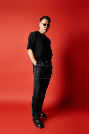 Foto de Un hombre asiático guapo y elegante con una camisa negra y pantalones negros en un ambiente de estudio, de pie con confianza sobre un fondo rojo. - Imagen libre de derechos