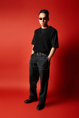 A stylish and handsome Asian man in black attire striking a pose in front of a vivid red background in a studio setting.