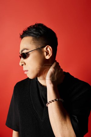 A handsome Asian man exudes style in a black shirt and sunglasses against a vibrant red background in a studio setting.