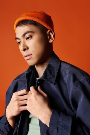 Handsome Asian man in blue jacket stands confidently in orange hat against vibrant studio backdrop.