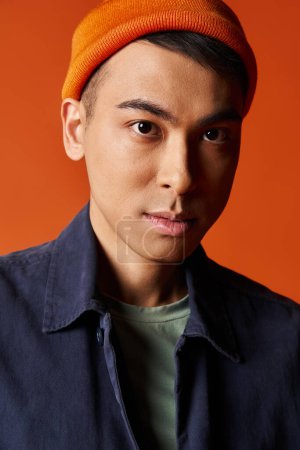A handsome Asian man in a stylish blue shirt and orange hat stands confidently against an orange background in a studio.