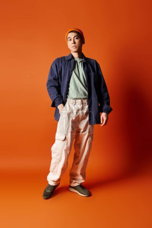 A handsome Asian man stands confidently in stylish attire against a vibrant orange background in a studio setting.