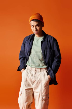 A handsome Asian man in a blue jacket and orange hat strikes a confident pose against an orange background in a studio.
